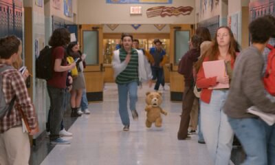 alt="El oso de peluche animado Ted y el joven John Bennett corren frenéticamente por el pasillo de un instituto de secundaria, en una escena de caos escolar de los años 90. John mira a la cámara con expresión asustada, mientras otros estudiantes se apartan. El fondo muestra casilleros azules y decoraciones escolares."