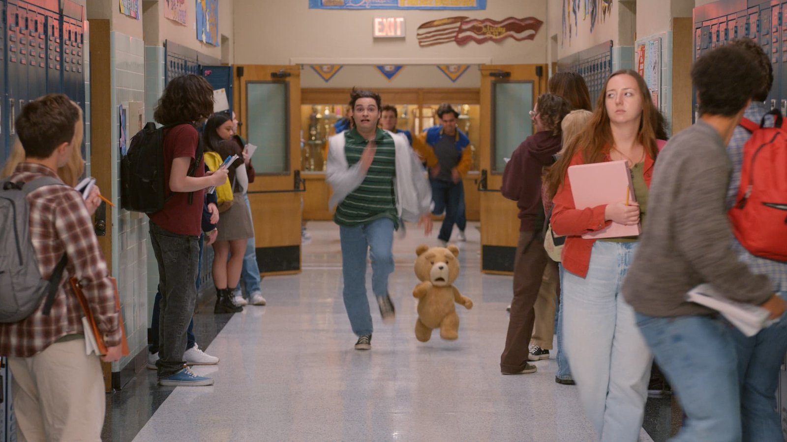 alt="El oso de peluche animado Ted y el joven John Bennett corren frenéticamente por el pasillo de un instituto de secundaria, en una escena de caos escolar de los años 90. John mira a la cámara con expresión asustada, mientras otros estudiantes se apartan. El fondo muestra casilleros azules y decoraciones escolares."
